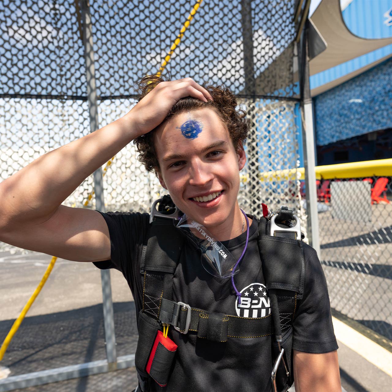 A newly licensed skydiver wearing his skydiving rig proudly pulls back his bangs to display his A license stamp on his forhead