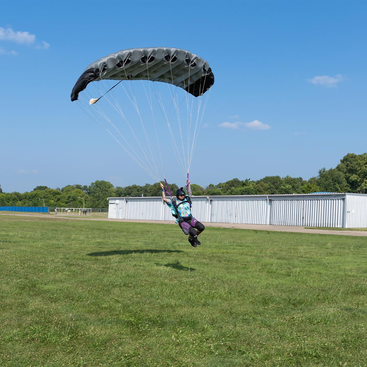 A licensed skydiver flying in for landing beneath a small gray and black parachute