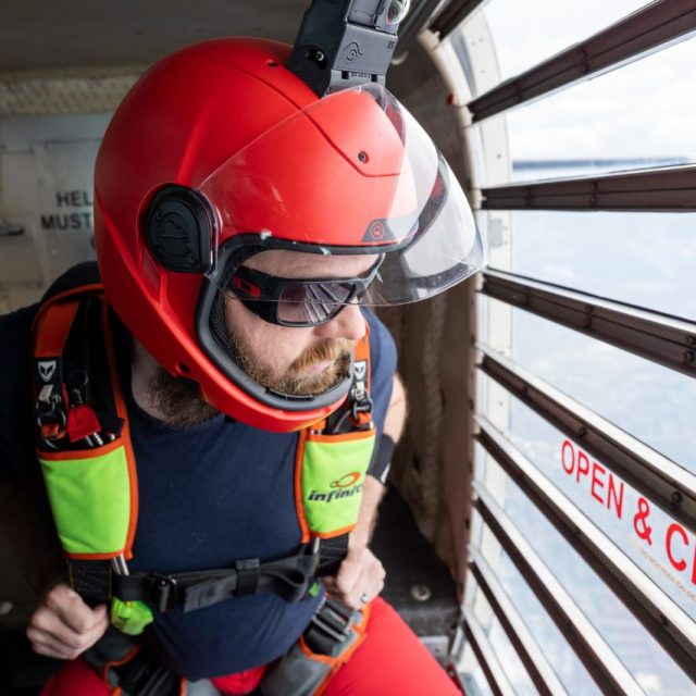 Male licensed jumper in a red helmet with a lime green and red skydiving rig gazes out the clear door of the airplane.