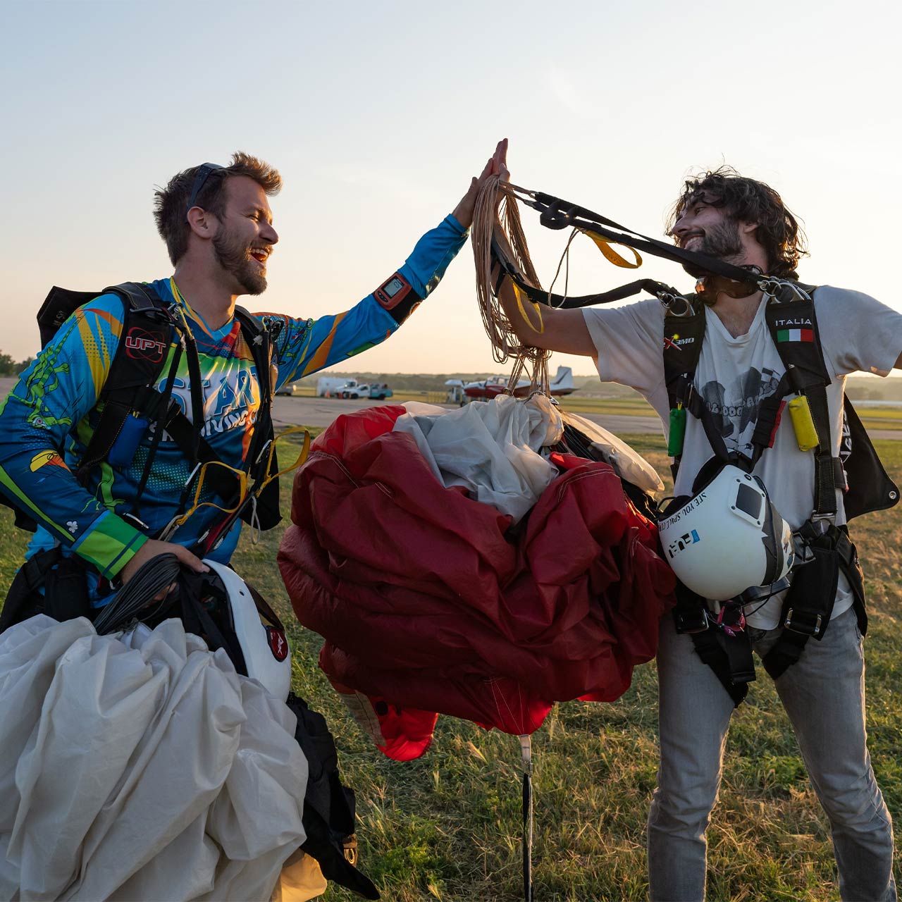 Two licensed skydivers holding their canopies high five jubilantly on the ground after landing