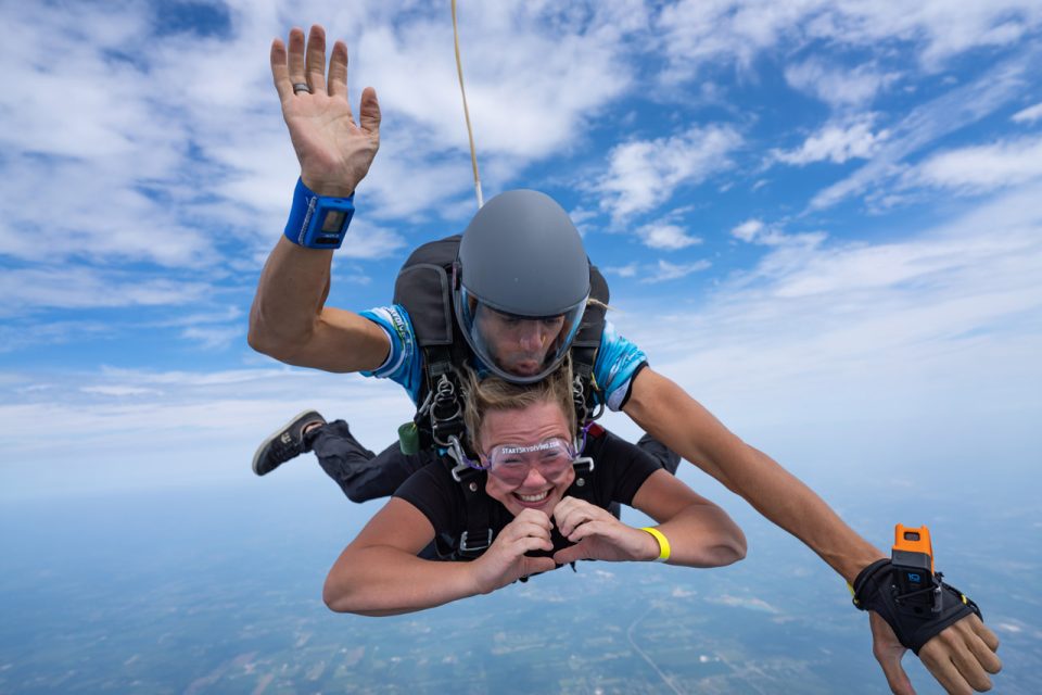Blonde female tandem student forming a heart with her hands as she freefalls on a tandem skydive at Start Skydiving