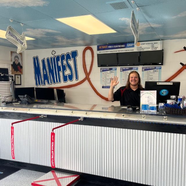A smiling worker stands waving behind the check-in desk at Start Skydiving