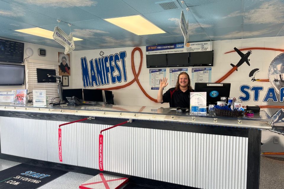 A smiling worker stands waving behind the check-in desk at Start Skydiving