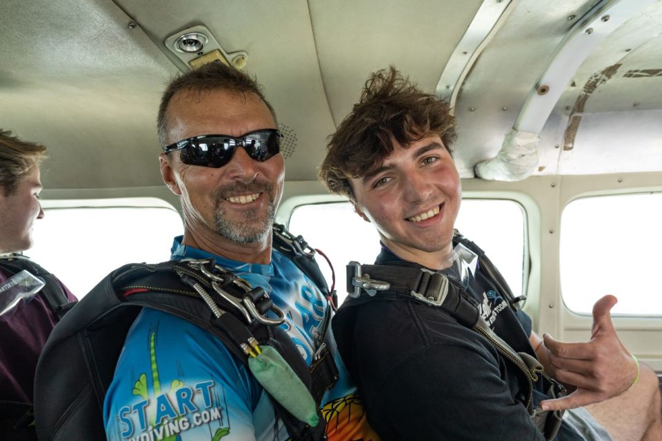 Male tandem skydiving pair with brown hair giving the shaka hand sign in the airplane on the way to altitude.