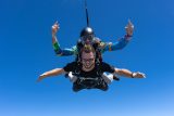 Male tandem skydiving pair wearing sunglasses in freefall with bright blue sky behind.