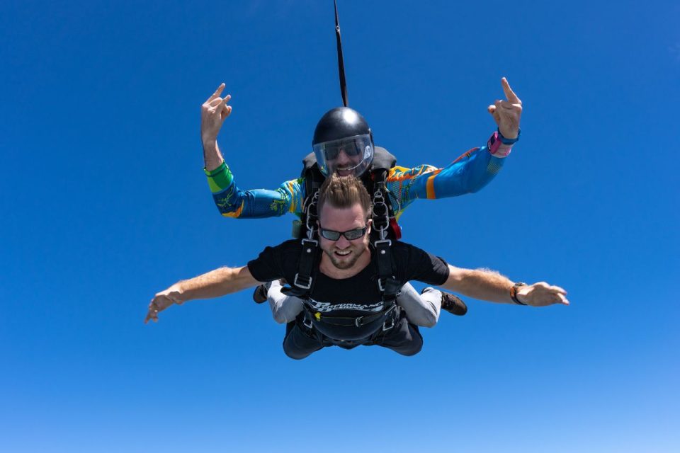 Male tandem skydiving pair wearing sunglasses in freefall with bright blue sky behind.