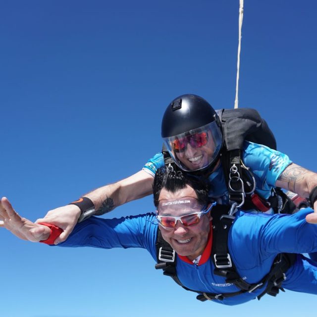 Smiling tandem instructor holds on to tandem students arms to help him extend them during freefall