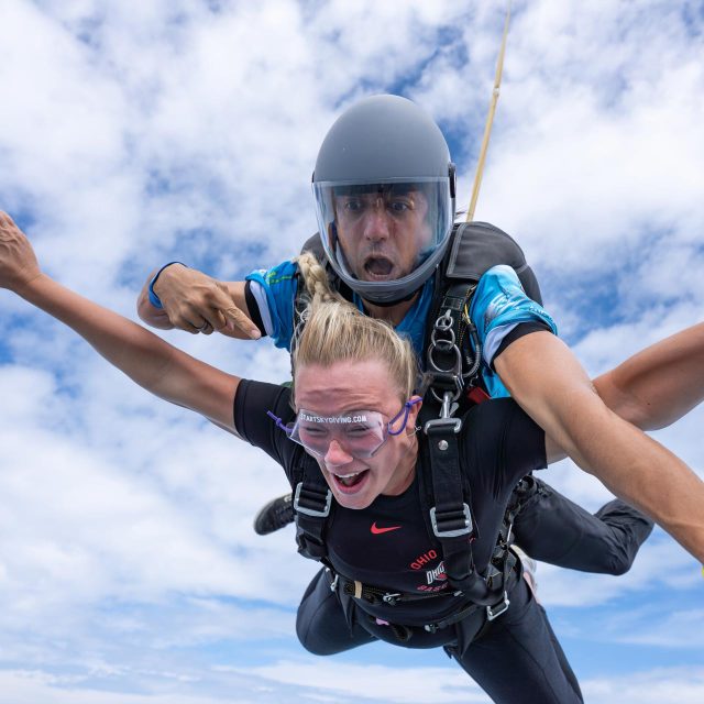 Tandem skydiving instructor points and blonde female skydiving student during freefall.