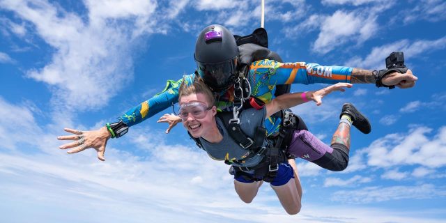 A male and female tandem skydiving pair with arms outstretched and a brilliant blue sky behind them.